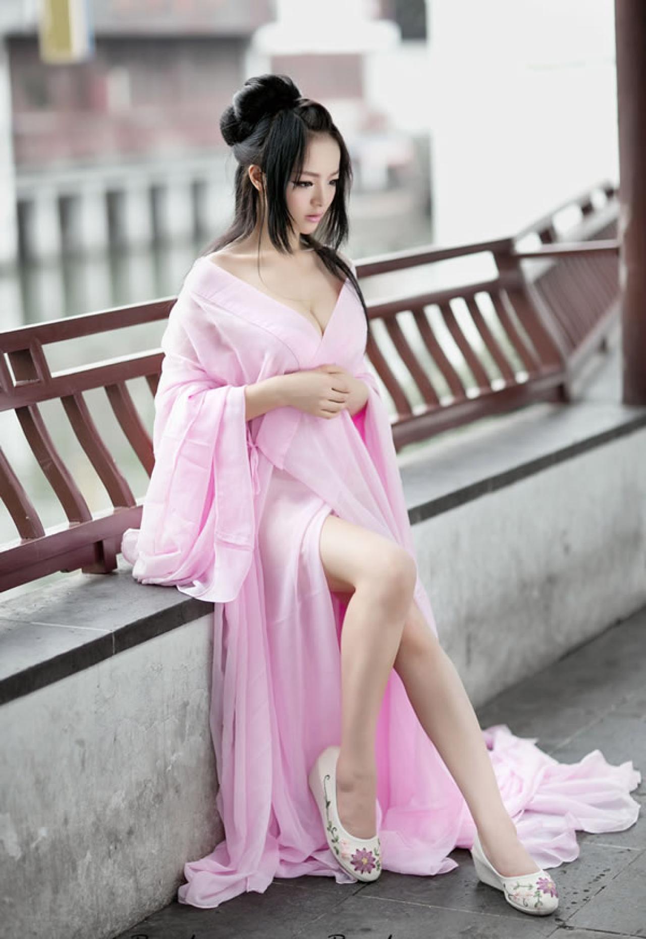 歪歪穿着粉色绸缎坐在木亭的长椅上双手放在胸下，低头略有所思