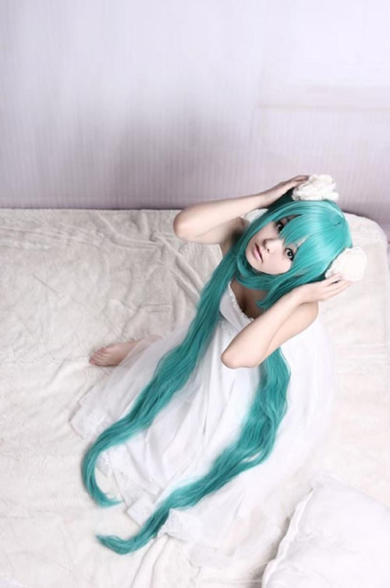 kuuya穿着白色睡衣扮演初音未来坐在床上，双手扶住双马尾的样子