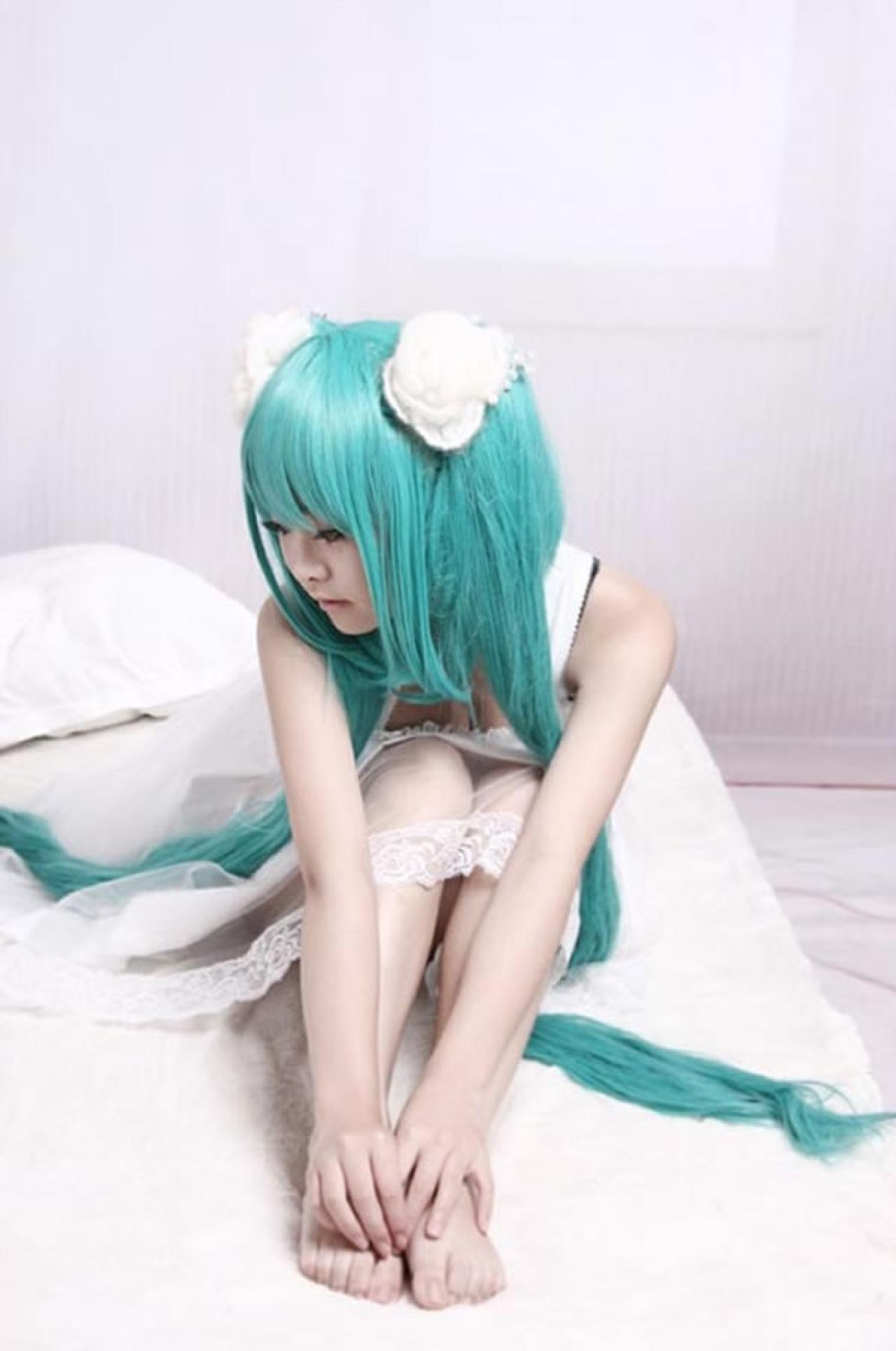 kuuya穿着白色睡衣扮演初音未来坐在床上，手摸到双脚，上半身蜷缩靠在腿上的正面