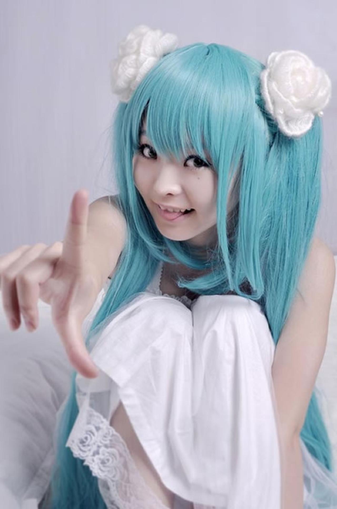 kuuya穿着白色睡衣扮演初音未来坐在床上，吐舌竖起食指，卖萌的样子