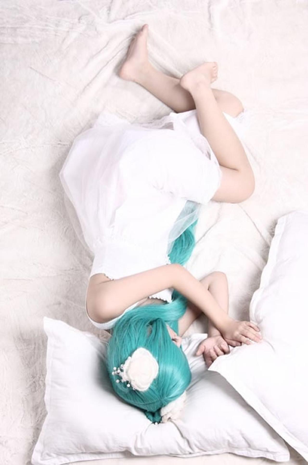 kuuya穿着白色睡衣扮演初音未来躺在白色枕头上，双手扶着另一个枕头，弯曲着大腿，睡着的样子