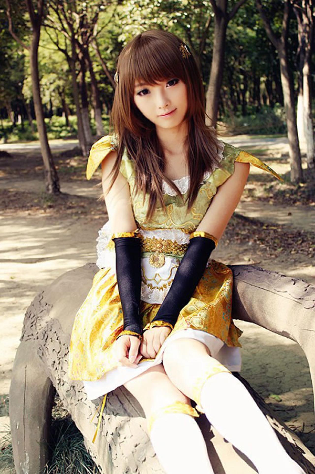 CC喵穿着黄色古装扮演小乔半坐在木头长椅上，歪着头微笑的样子