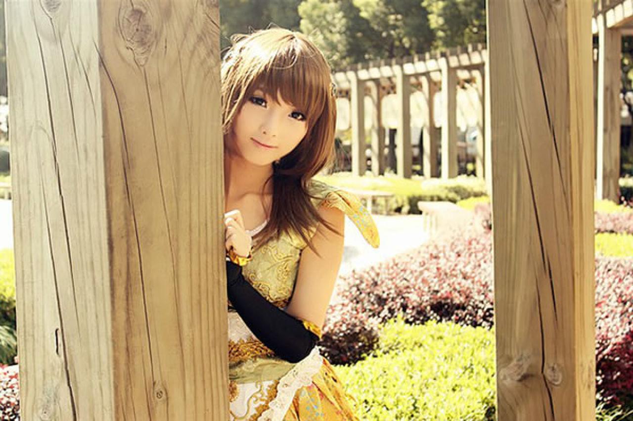 CC喵穿着黄色古装扮演小乔在一个长廊中，藏在一个树皮纹理的柱子后面，露出头，一手扶着柱子