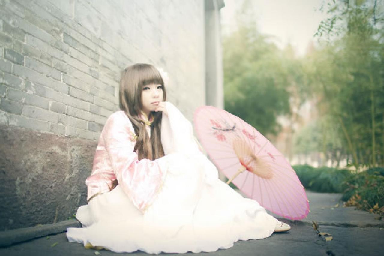 季_凋葬穿着粉衣扮演湾娘靠坐在老城墙上，嘟嘴轻咬着手指，粉色油纸伞放在身旁