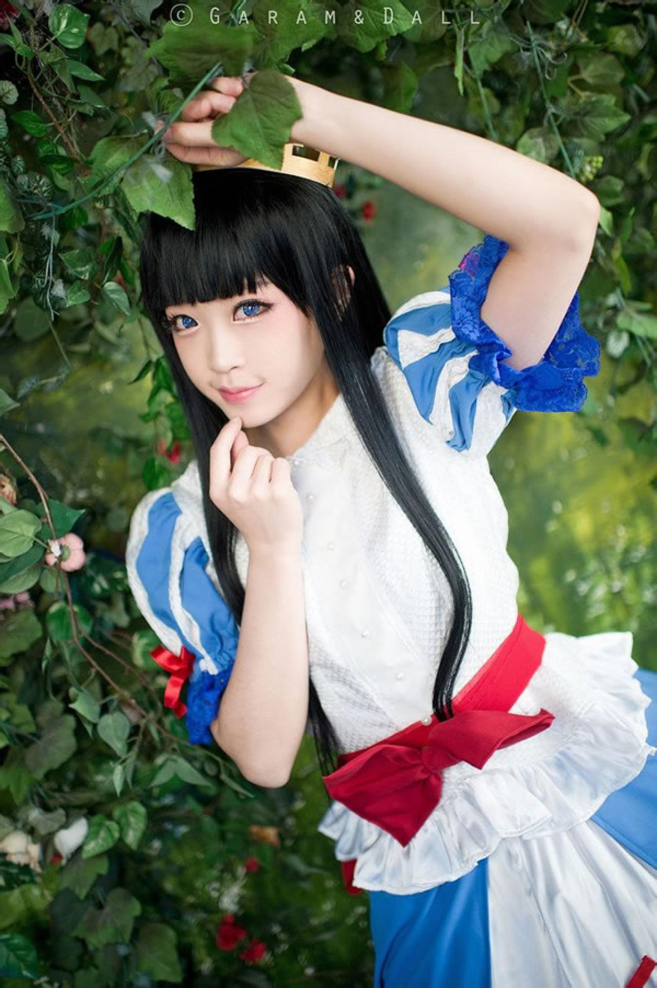 兔娘Tomia穿着蓝白洋装扮演白雪公主站在葡萄滕旁一手拽着葡萄藤叶，另一只手抵住下吧，微笑的样子