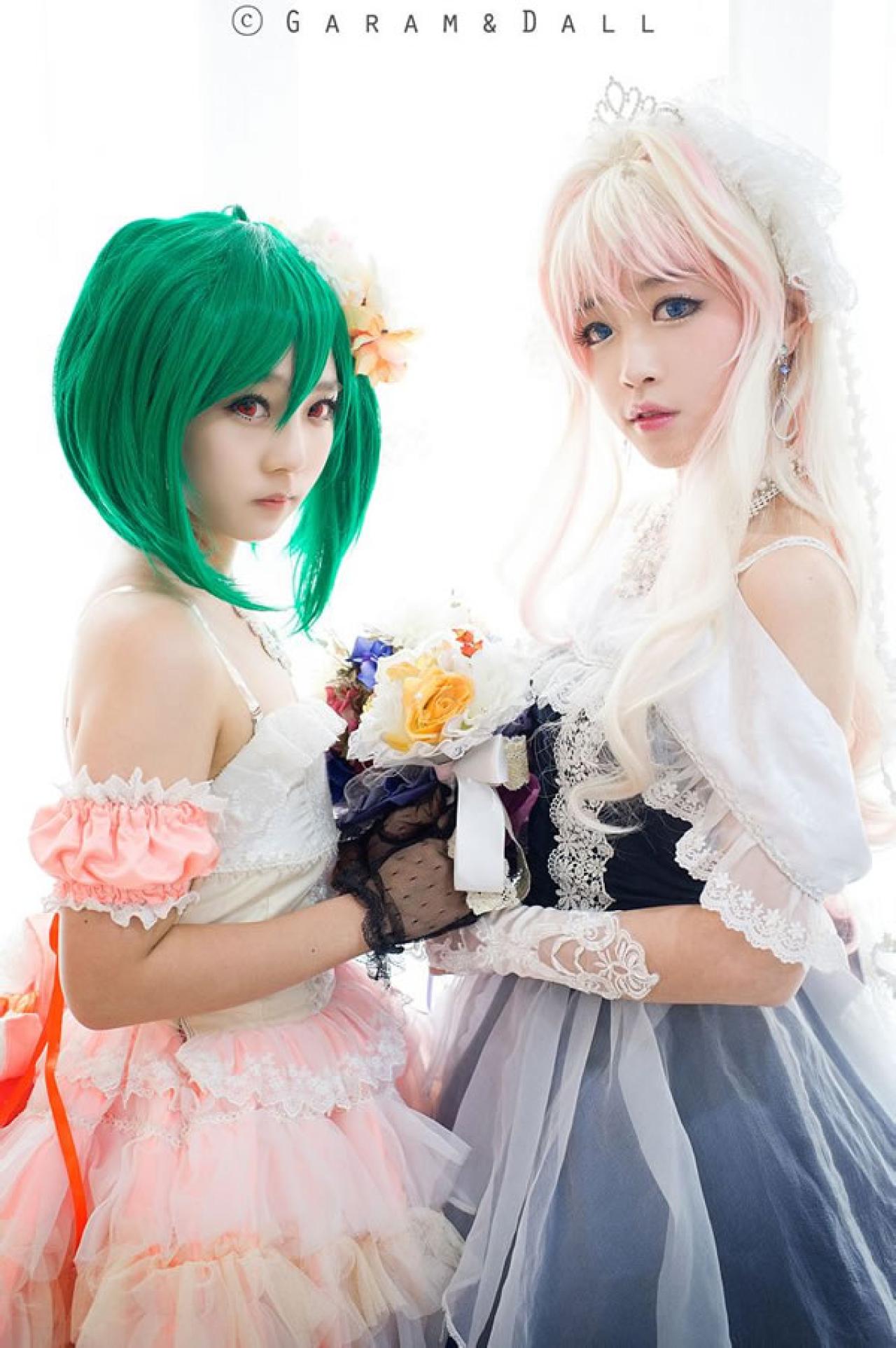 雪露·诺姆穿着蓝色洋装提着裙摆站在穿着粉色洋装的兰歌·李两人颔首捧着一束鲜花面对面，脸都看向镜头