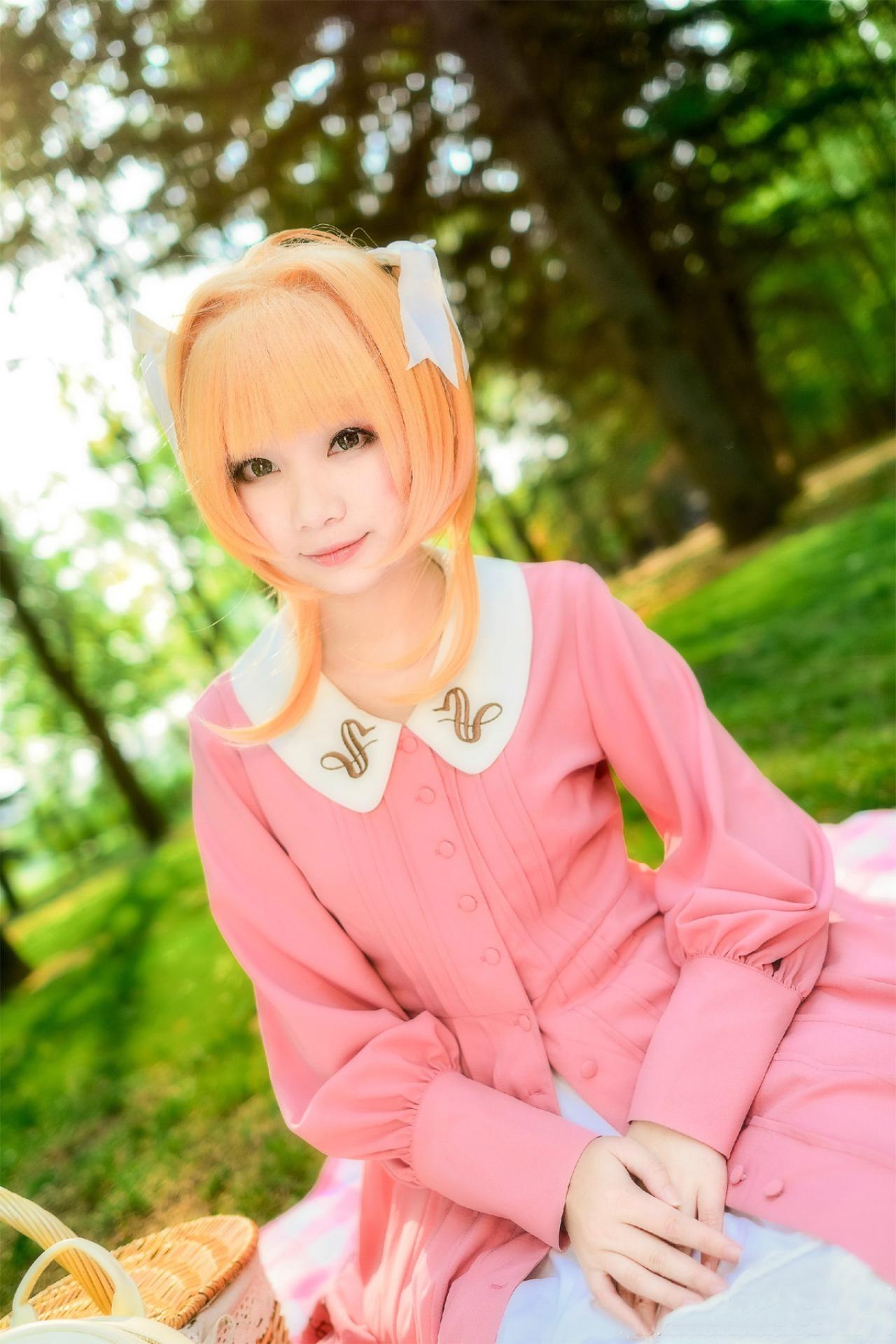 绯爱酱扮演木之本樱穿着粉色上衣与白色长裙坐在草地上微笑的样子