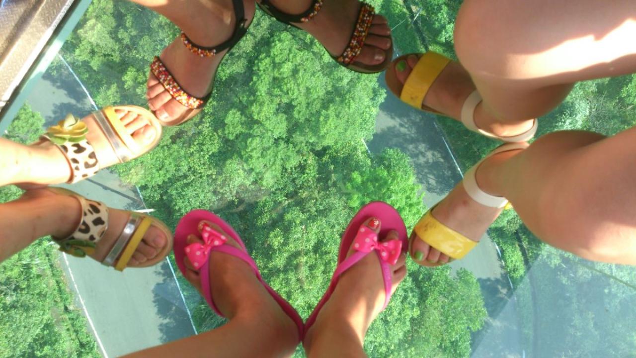 修老虎和小伙伴站在透明的缆车上，大伙脚撇成八字，靠在一起呈四边形，俯拍各自穿着凉鞋的脚的照片