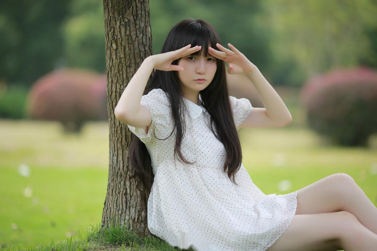 苏里柟穿着白色波点罩衫靠在树上，双手放在眼睛上方，遮挡阳光，正视镜头