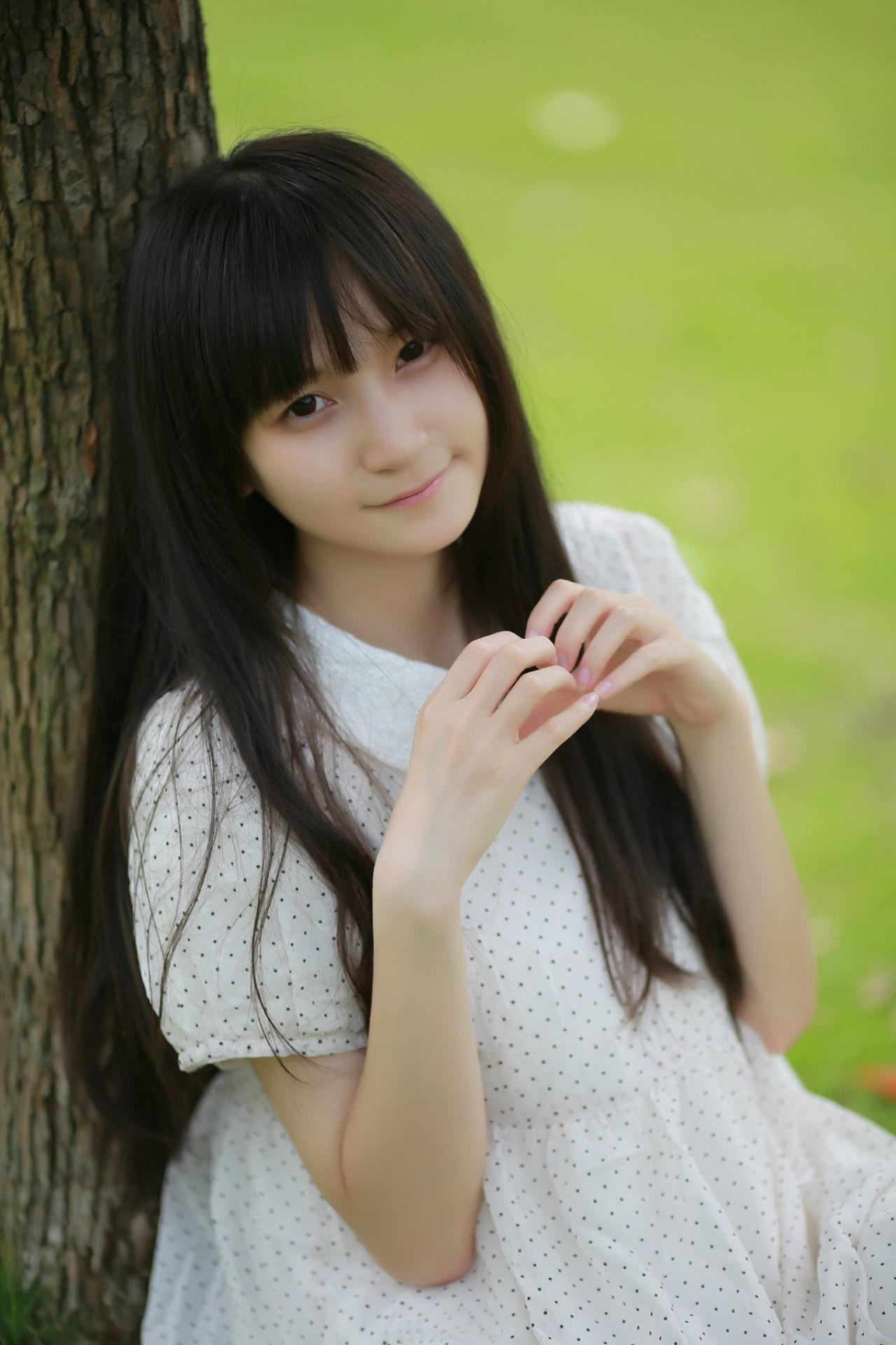 苏里柟穿着白色波点罩衫靠在树上，作心形手势
