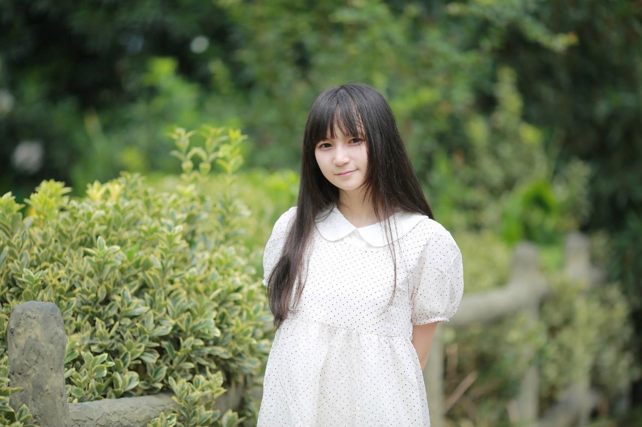 苏里柟穿着白色波点罩衫手背后站在篱笆旁