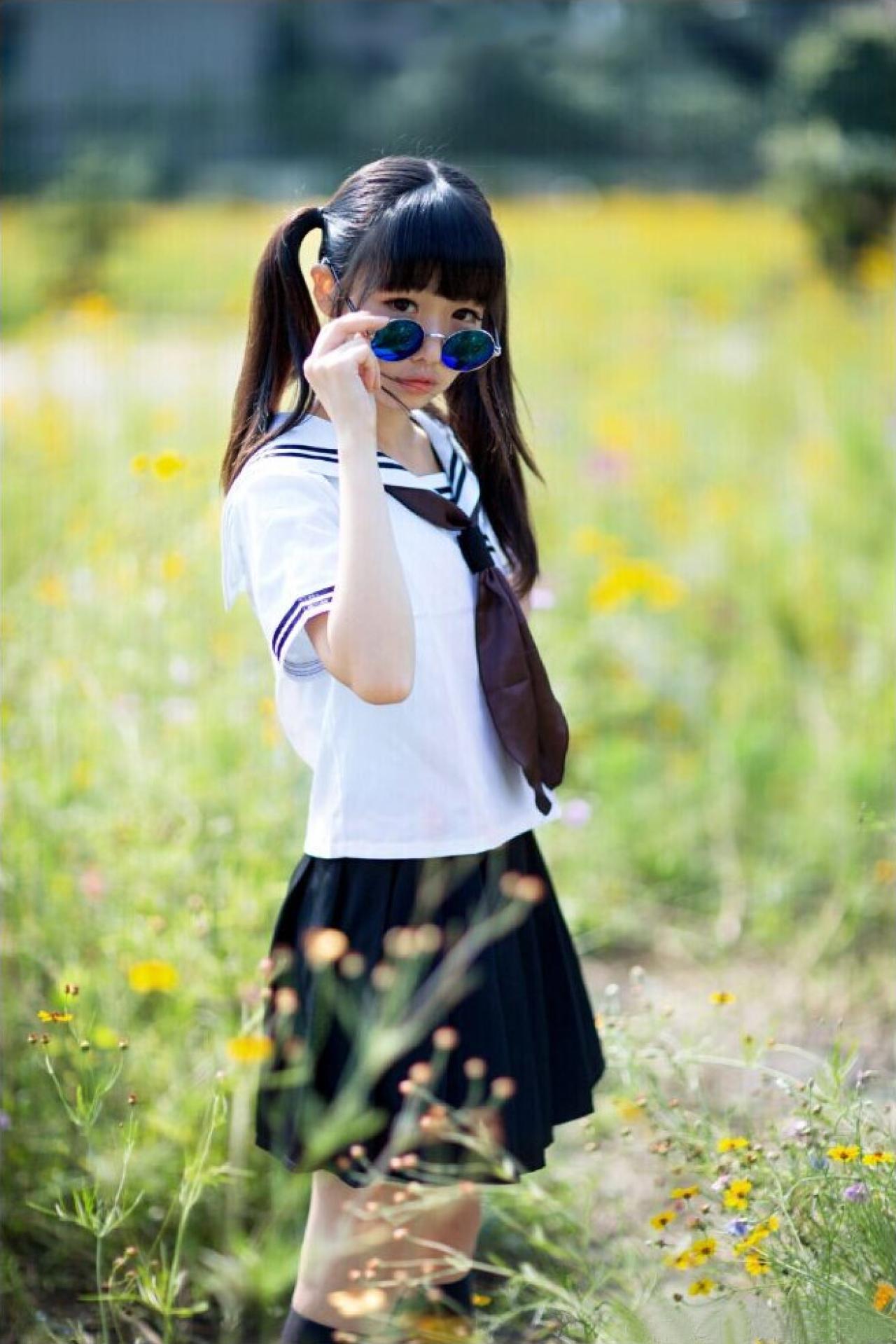 草纸戴着眼镜正在黄花碧草的田野之中，身体微侧站立，略微扶了一下眼镜