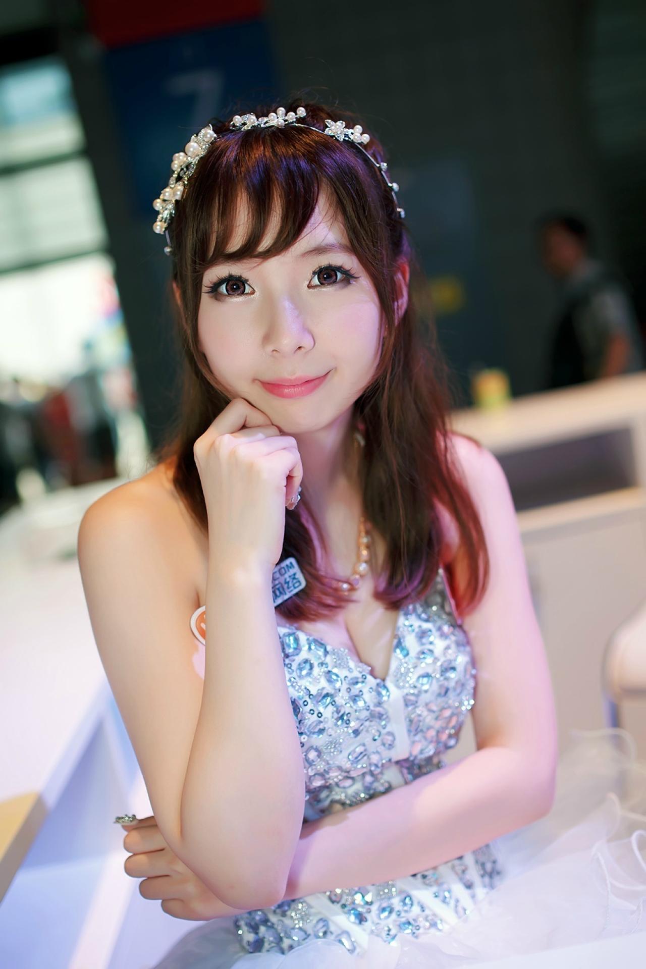 穿着布满水晶饰片白色连衣裙的XX网络Showgirl正坐在展台，左手放在腰间，右手半握拳抵住下巴