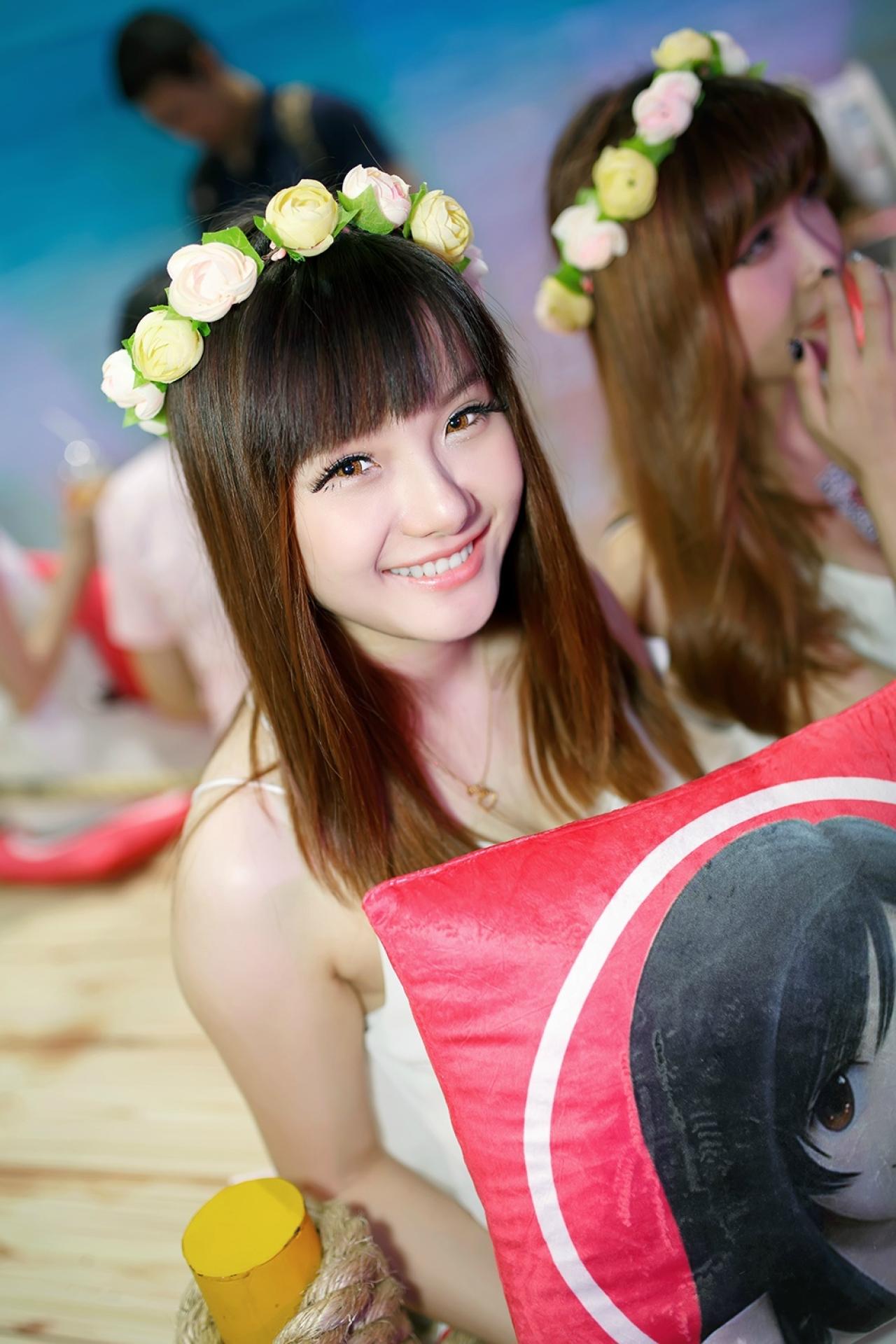 穿着白色连衣裙，头戴花环的Showgirl拿着印有百思不得姐Logo的抱枕对着镜头咧嘴微笑