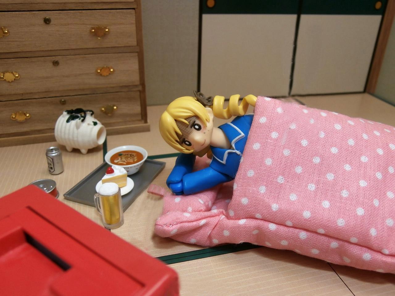 巴麻美的手办穿着蓝色校服一脸黑线地躺在被窝里，旁边放着面和点心