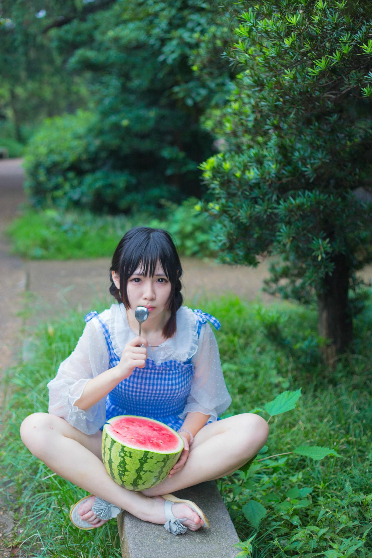 九樱喵盘腿坐在石凳上，将西瓜放在腿上，左手扶着西瓜，右手将小勺放在嘴边的姿势。