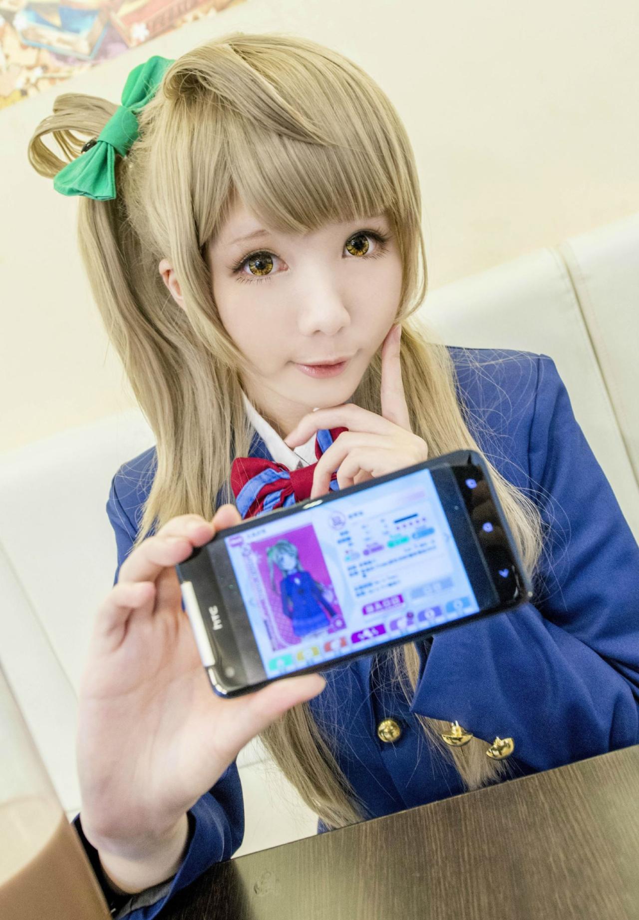 霜月扮演南小鸟穿着蓝色支付，左手戳脸卖萌，右手端着手机展示LoveLive!游戏中同样装扮的南小鸟卡牌