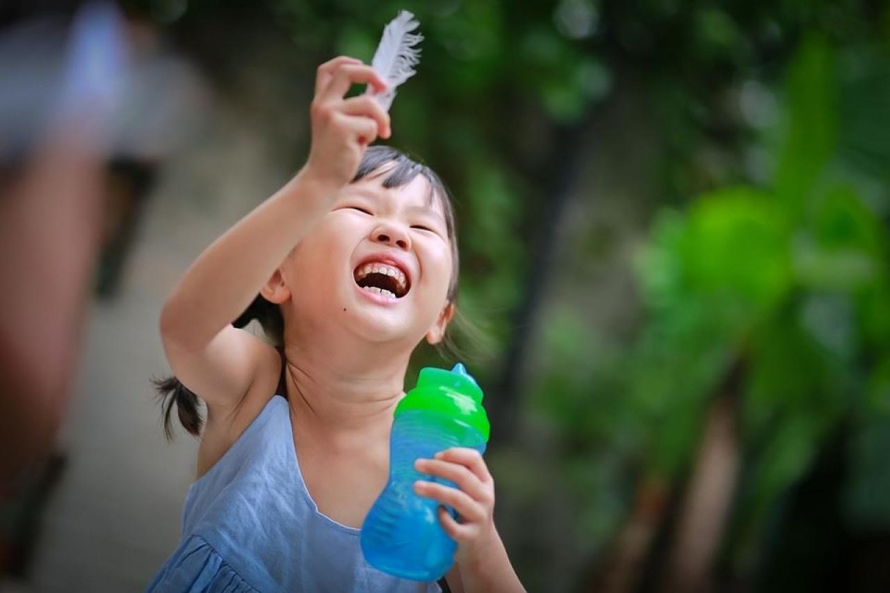 小女孩右手拿着羽毛，左手抱着水杯，开怀大笑的样子