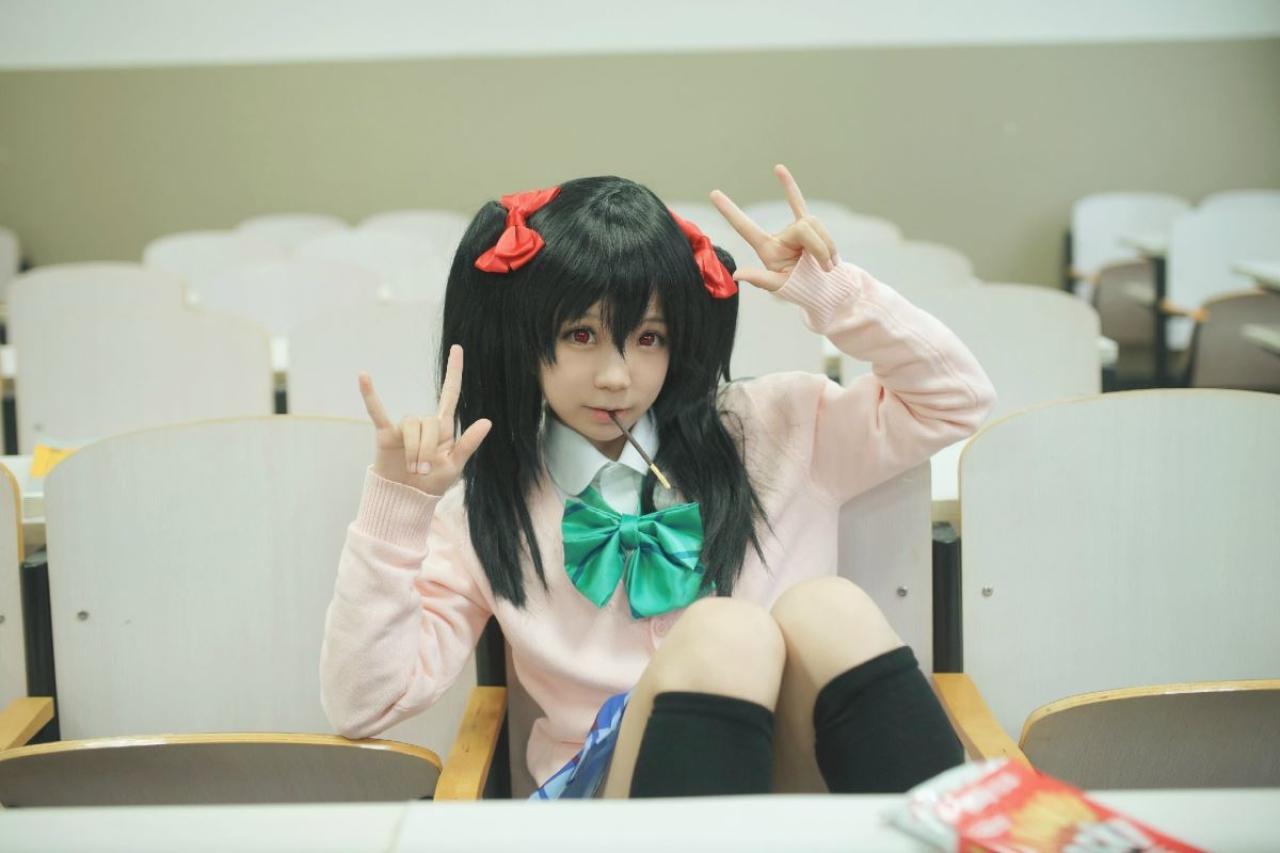 阿琴扮演矢泽妮可坐在阶梯教室的椅子上，翘着腿，叼着铅笔，双手作ILOVEYOU手势