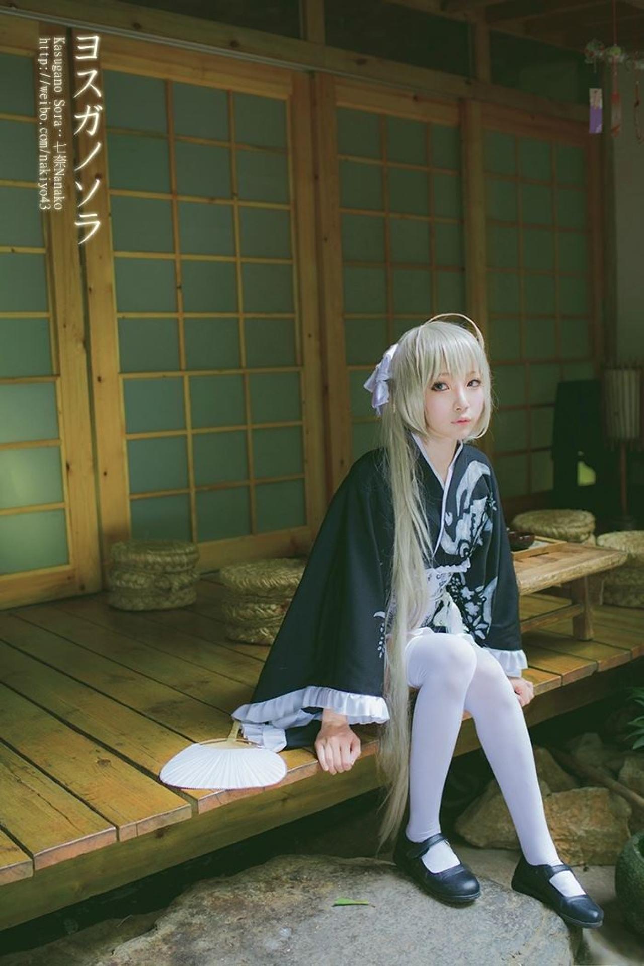 七奈扮演春日野穹坐在门前，上身前伸，身体一旁摆着扇子