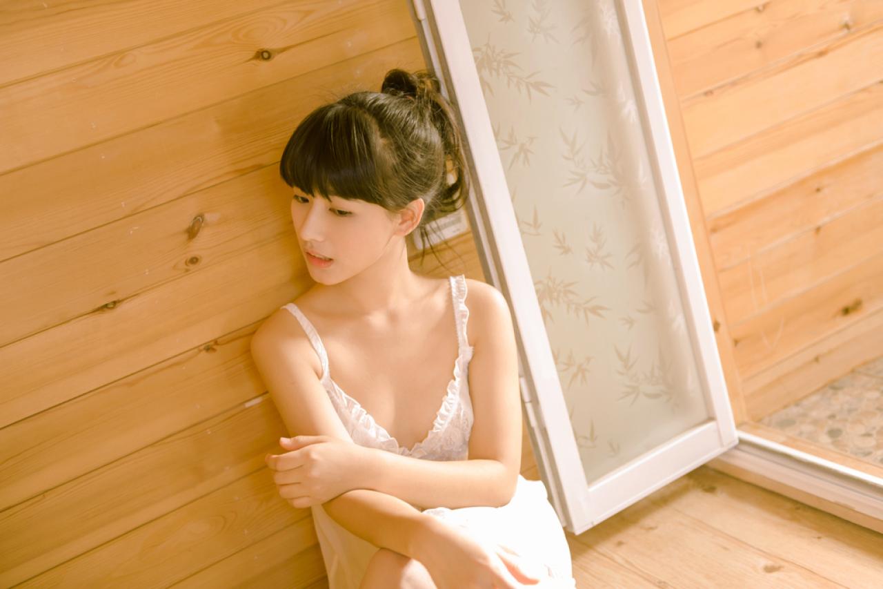 萌宅女靠在木纹墙坐在地板上，双臂放在腿上，歪着头