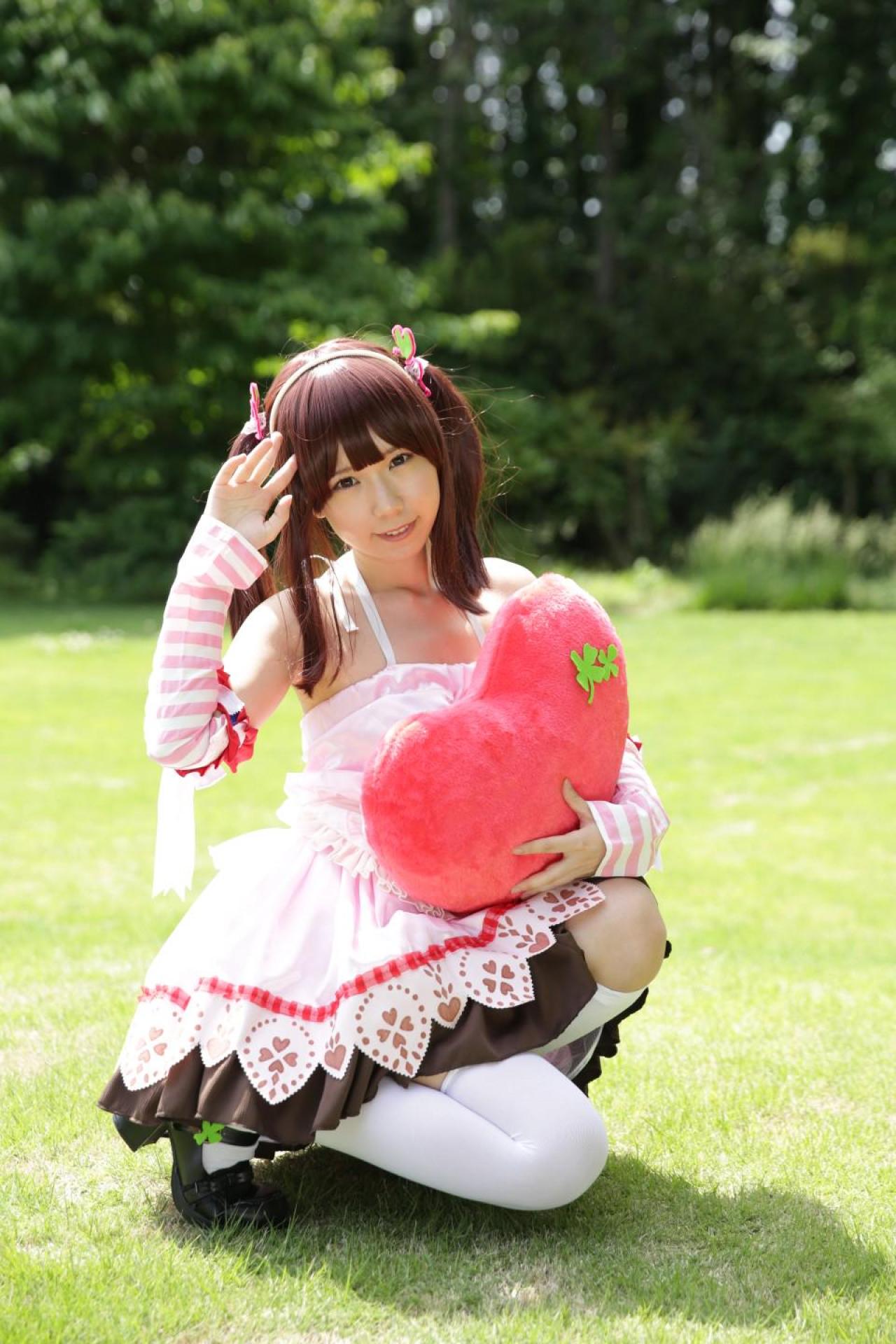 Reco扮绪方智绘里左手捧着桃心抱枕，右手放在额头右侧，蹲在草坪上