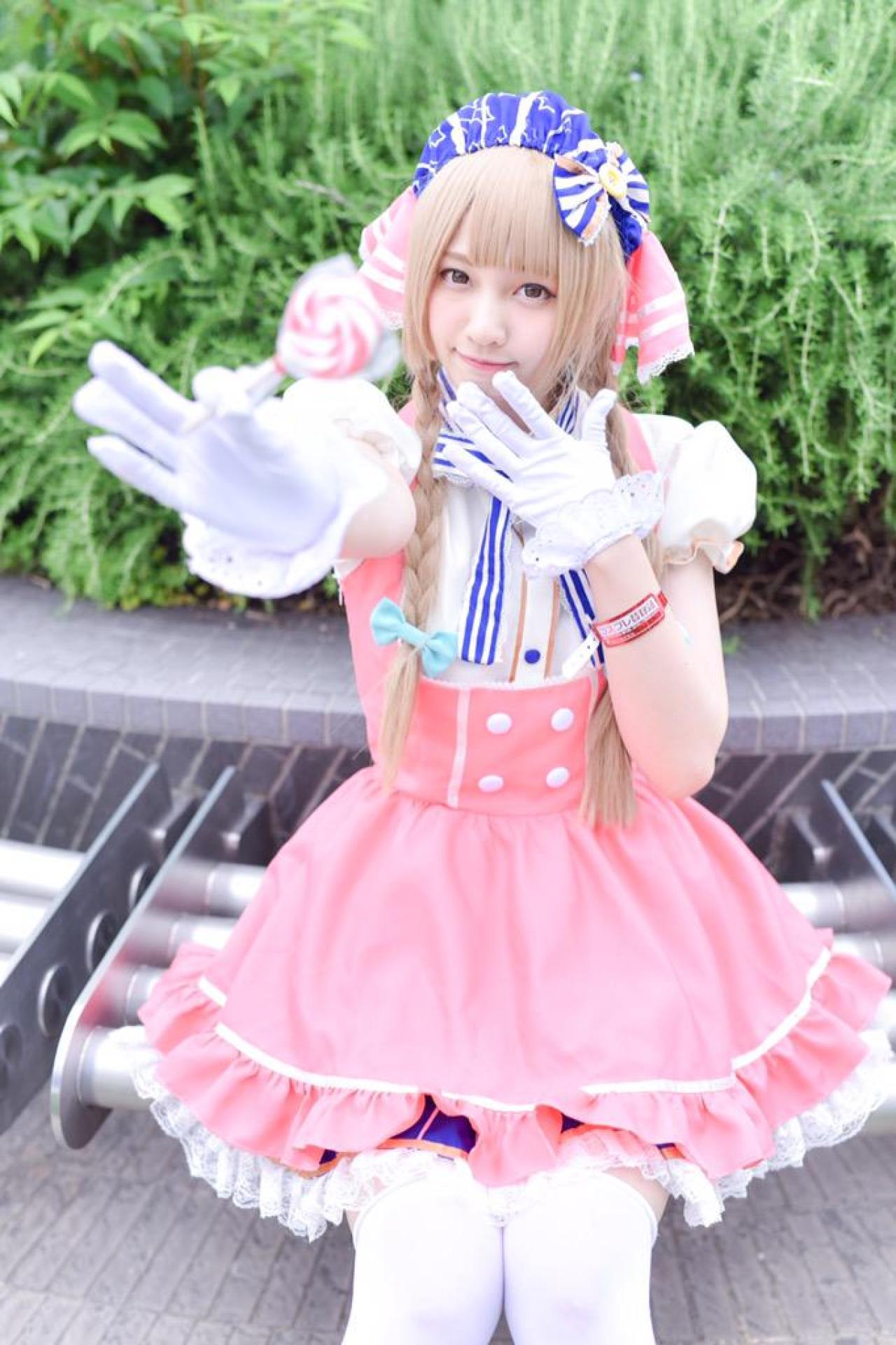 玖音さん扮南小鸟右手拿着棒棒糖，左手轻扶下巴坐在花池前