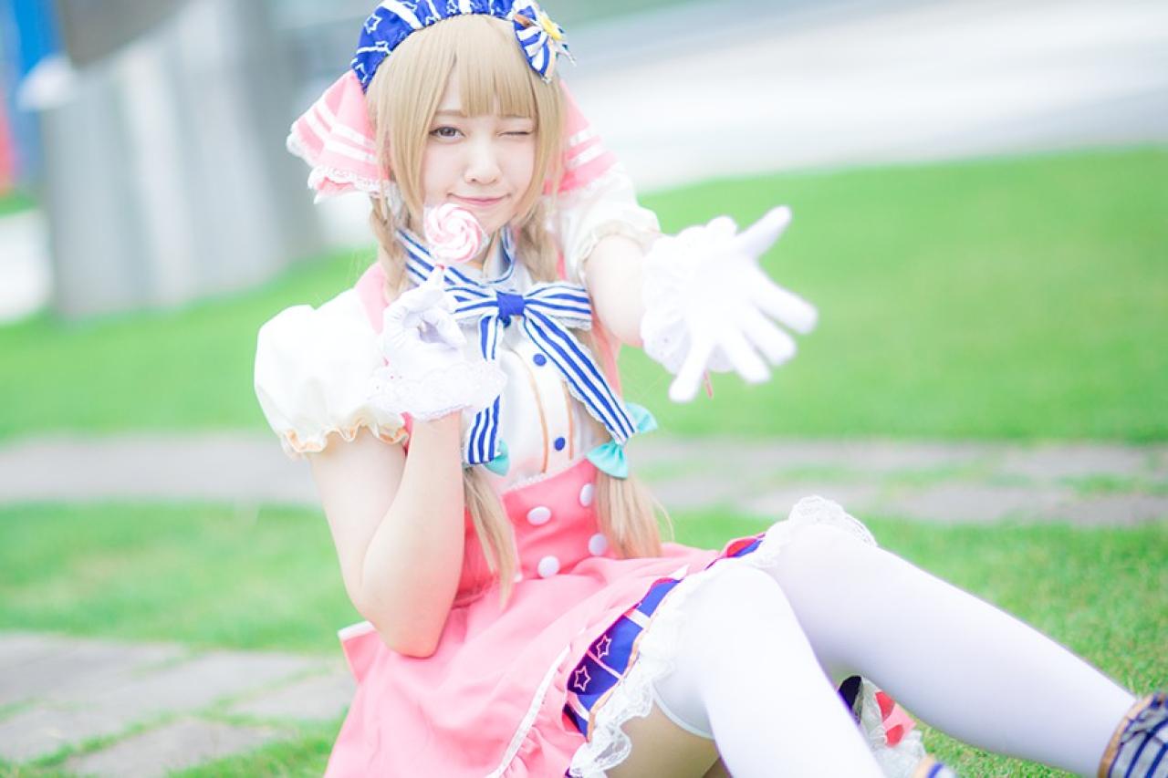 玖音さん扮南小鸟坐在草地，右手拿着棒棒糖，左手五指张开，眨眼卖萌