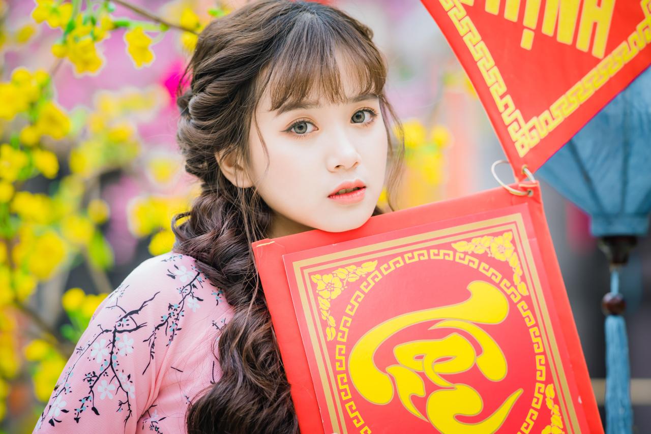 棕色双马尾卷发，齐刘海儿，穿着粉色旗袍的越南少女 站姿 脸部特写 正面照 横幅 双手抱着红色福字中国结 微笑 腊梅 黄梅 树