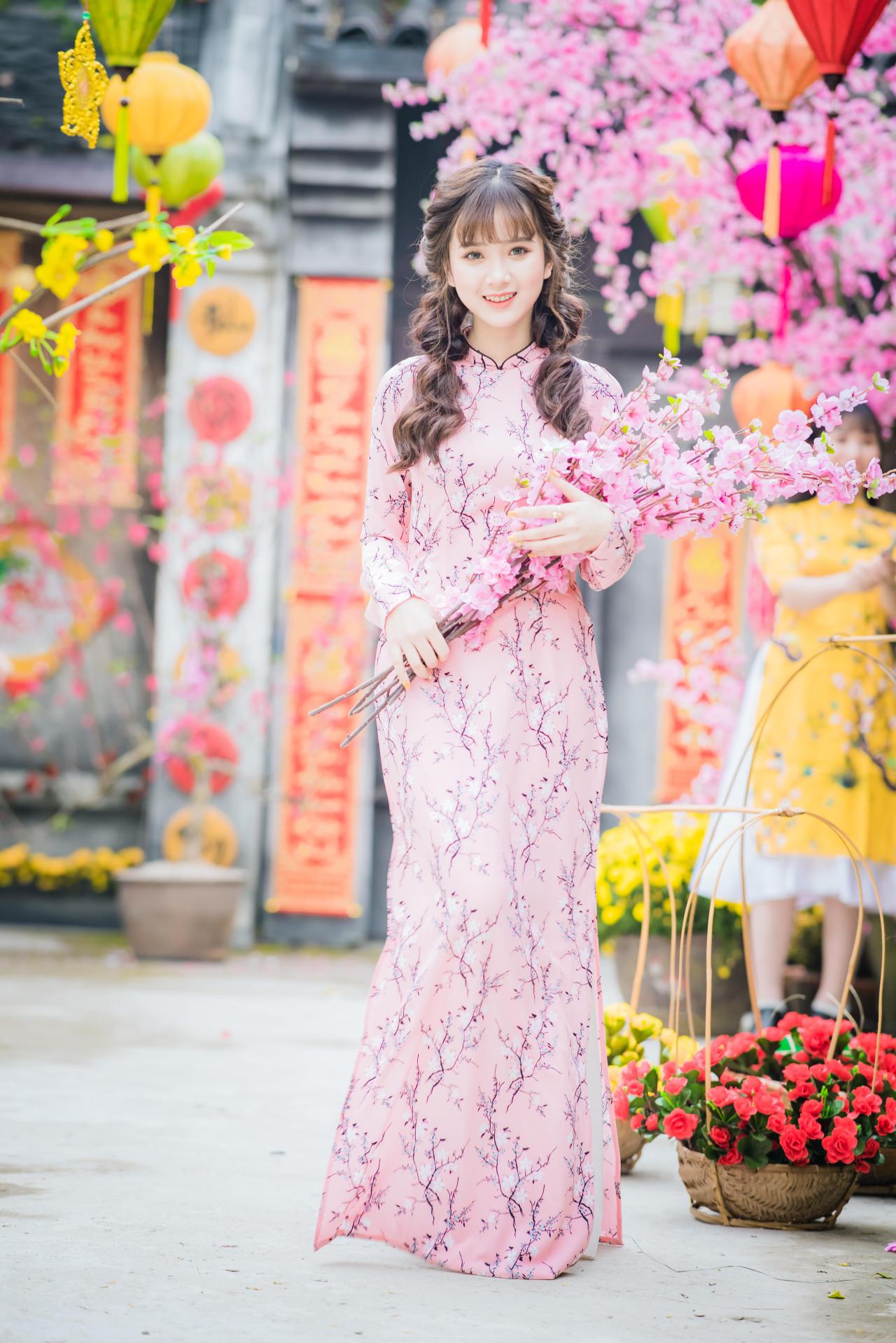 棕色双马尾卷发，齐刘海儿，穿着粉色旗袍的越南少女 站姿 全身照 正面照 竖幅 双手捧着一束粉色樱花 咧嘴露齿微笑 腊梅 黄梅 花篮 灯笼