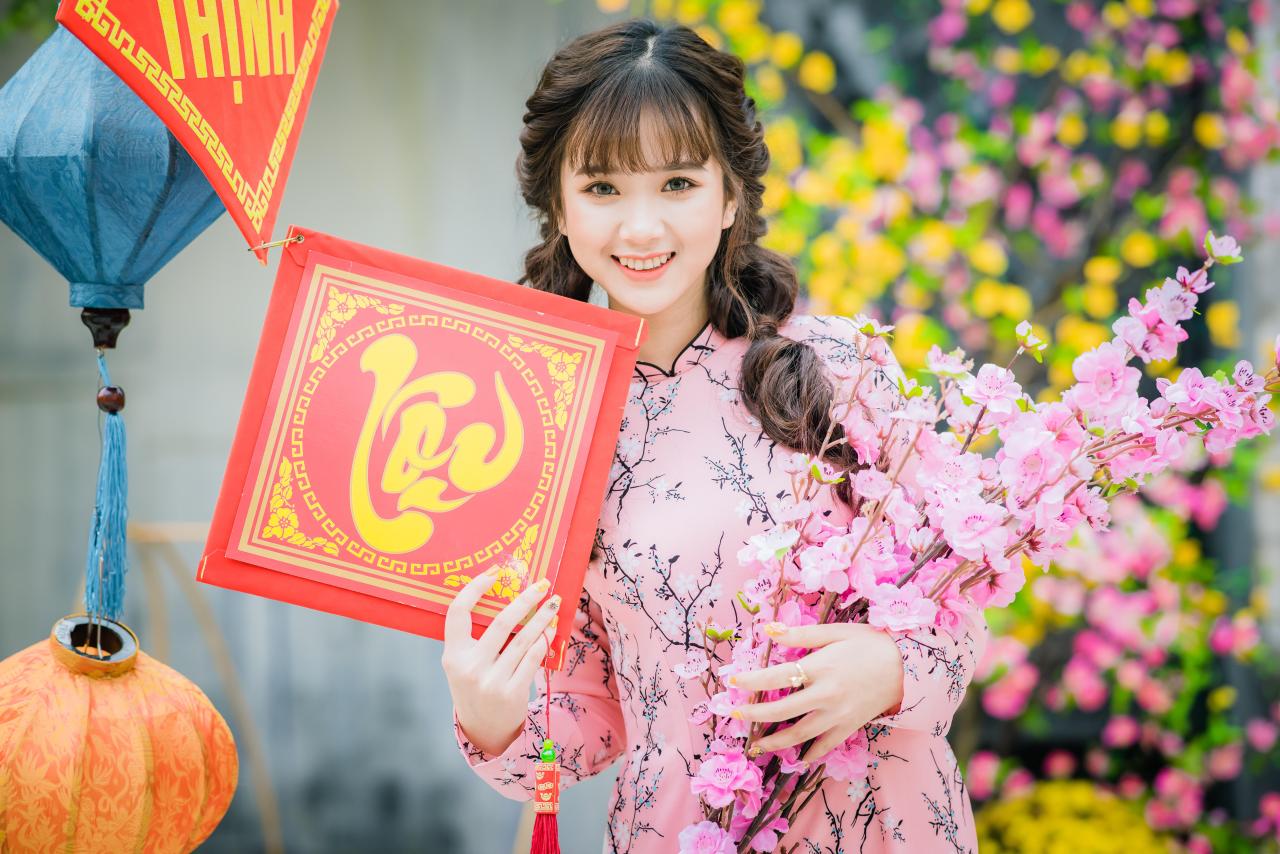 棕色双马尾卷发，齐刘海儿，穿着粉色旗袍的越南少女 站姿 上半身照 正面照 横幅 咧嘴露齿微笑 右手拿着福字中国结 左手捧着一束粉色樱花 腊梅 黄梅 花篮 蓝色和橙色灯笼