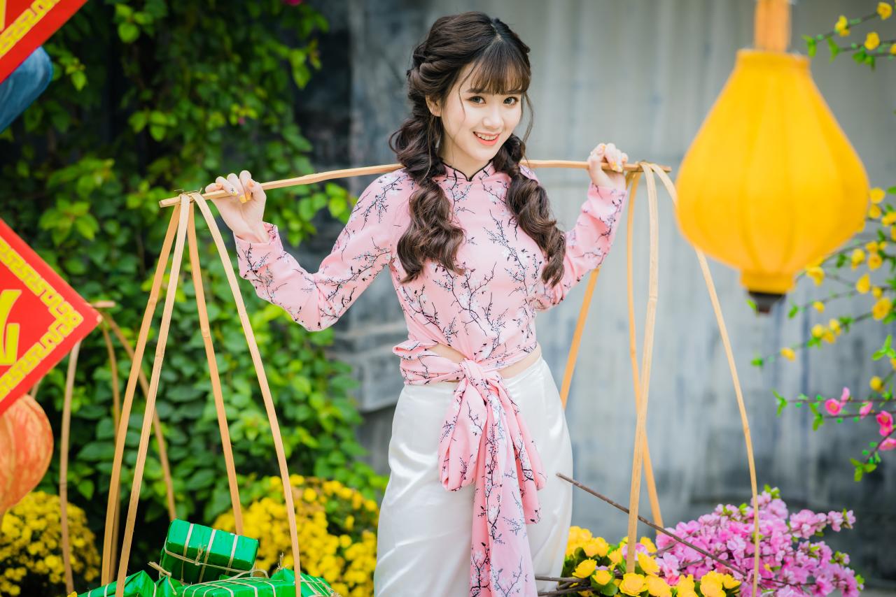 棕色双马尾卷发，齐刘海儿，穿着粉色旗袍白色长裤的少女 站姿 上半身照 正面照 横幅 咧嘴露齿微笑 扛着扁担挑着花篮 双手握住扁担 腊梅 黄梅 花篮 蓝色和黄色灯笼 绿色年货包