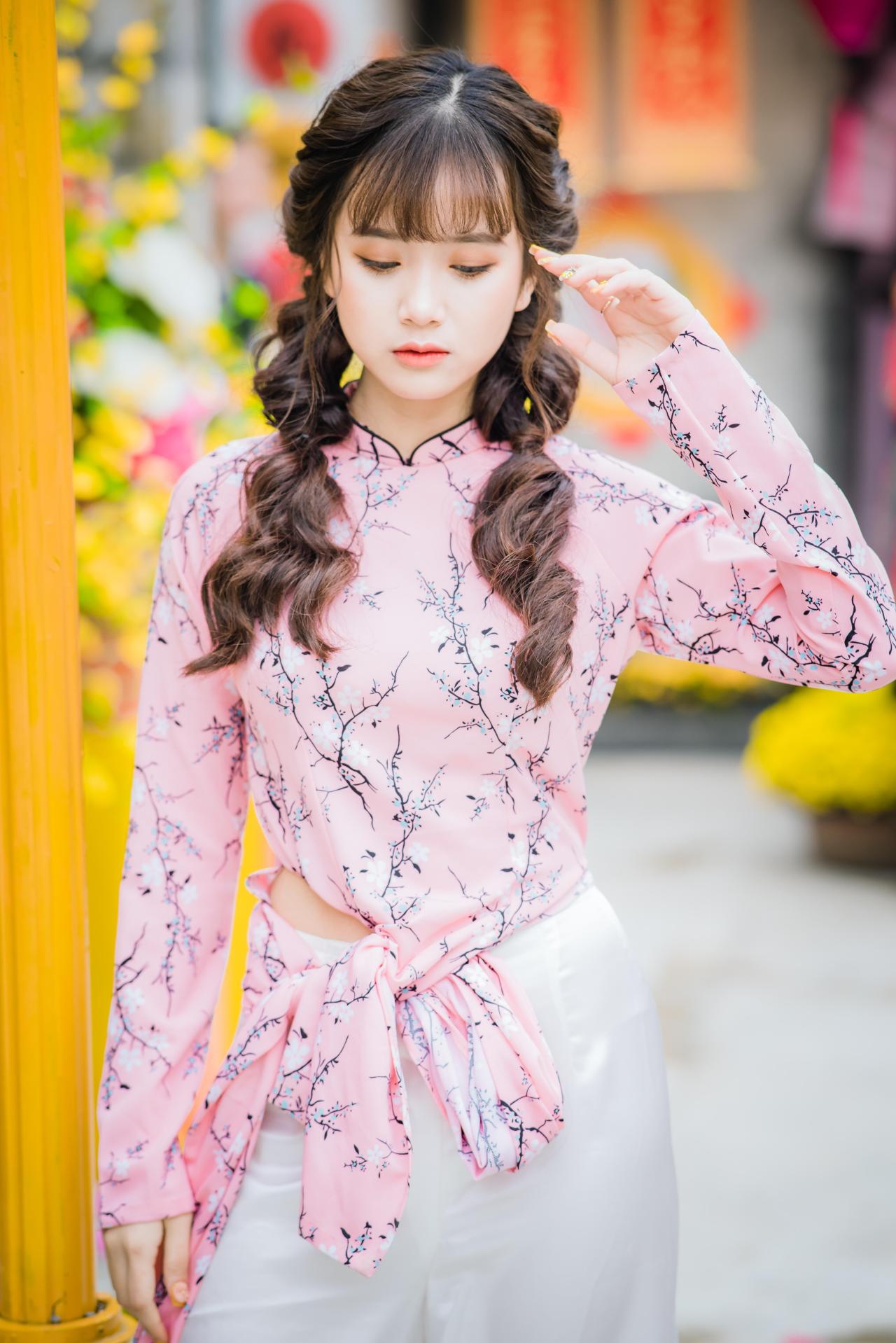 棕色双马尾卷发，齐刘海儿，穿着粉色旗袍白色长裤的少女 站姿 全身照 正面照 低头 左手手指指着额头 橙色灯柱