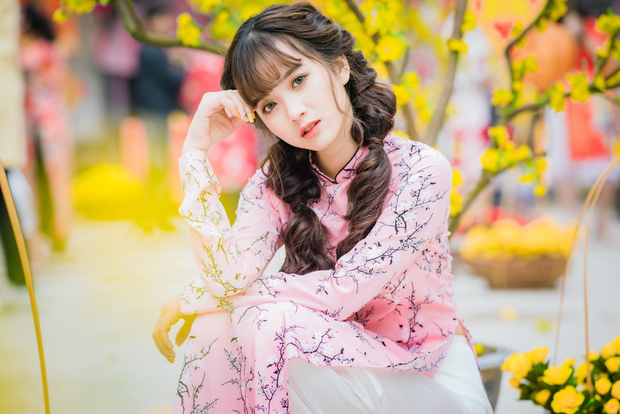 棕色双马尾卷发，齐刘海儿，穿着粉色旗袍的越南少女 坐姿 上半身照 4/3侧面照 横幅 右手轻抚额头 思考 腊梅 黄梅 花篮