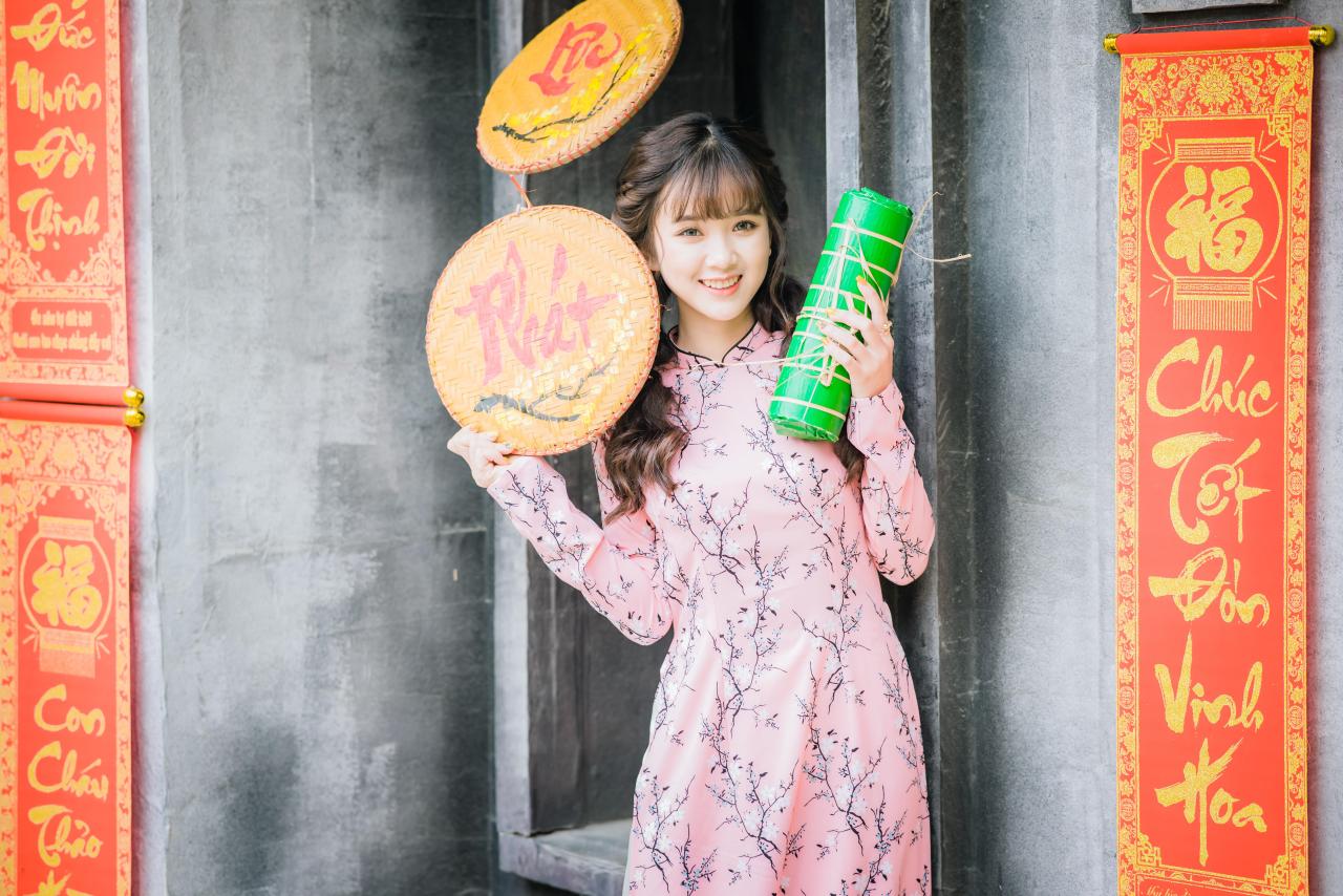 棕色双马尾卷发，齐刘海儿，穿着粉色旗袍白色长裤的少女 站姿 上半身照 正面照 横幅 咧嘴露齿微笑 左手拿着绿色年货包 右手拿着越南草帽 越南福字春联