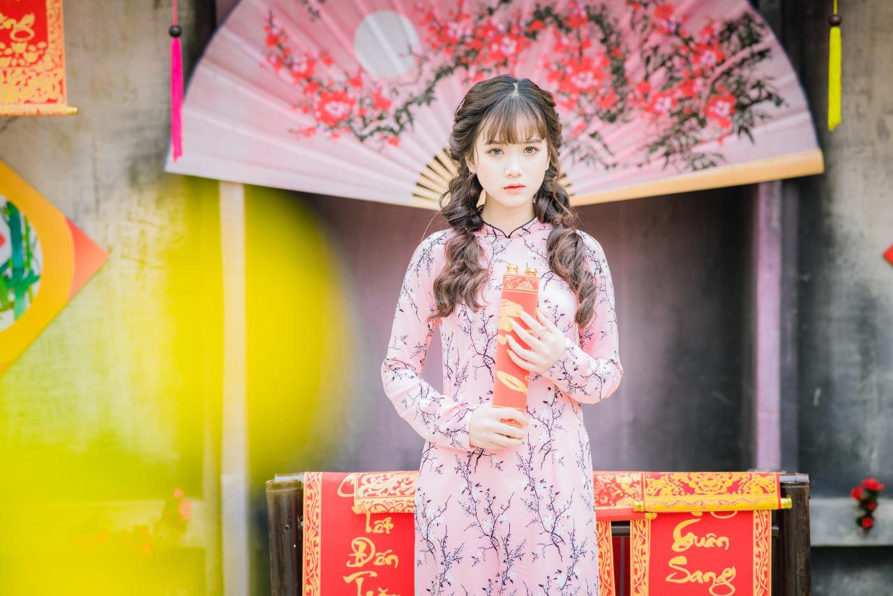 棕色双马尾卷发，齐刘海儿，穿着粉色旗袍白色长裤的少女 站姿 上半身照 正面照 横幅 双手捧着卷起的越南福字春联 粉色梅花扇