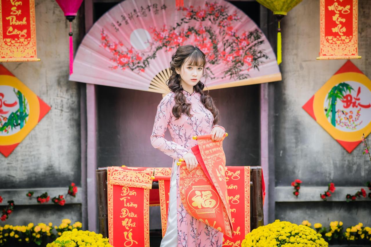 棕色双马尾卷发，齐刘海儿，穿着粉色旗袍白色长裤的少女 站姿 4/3侧面照 正面照 横幅 咧嘴露齿微笑 双手拿着打开的越南福字春联 粉色梅花扇