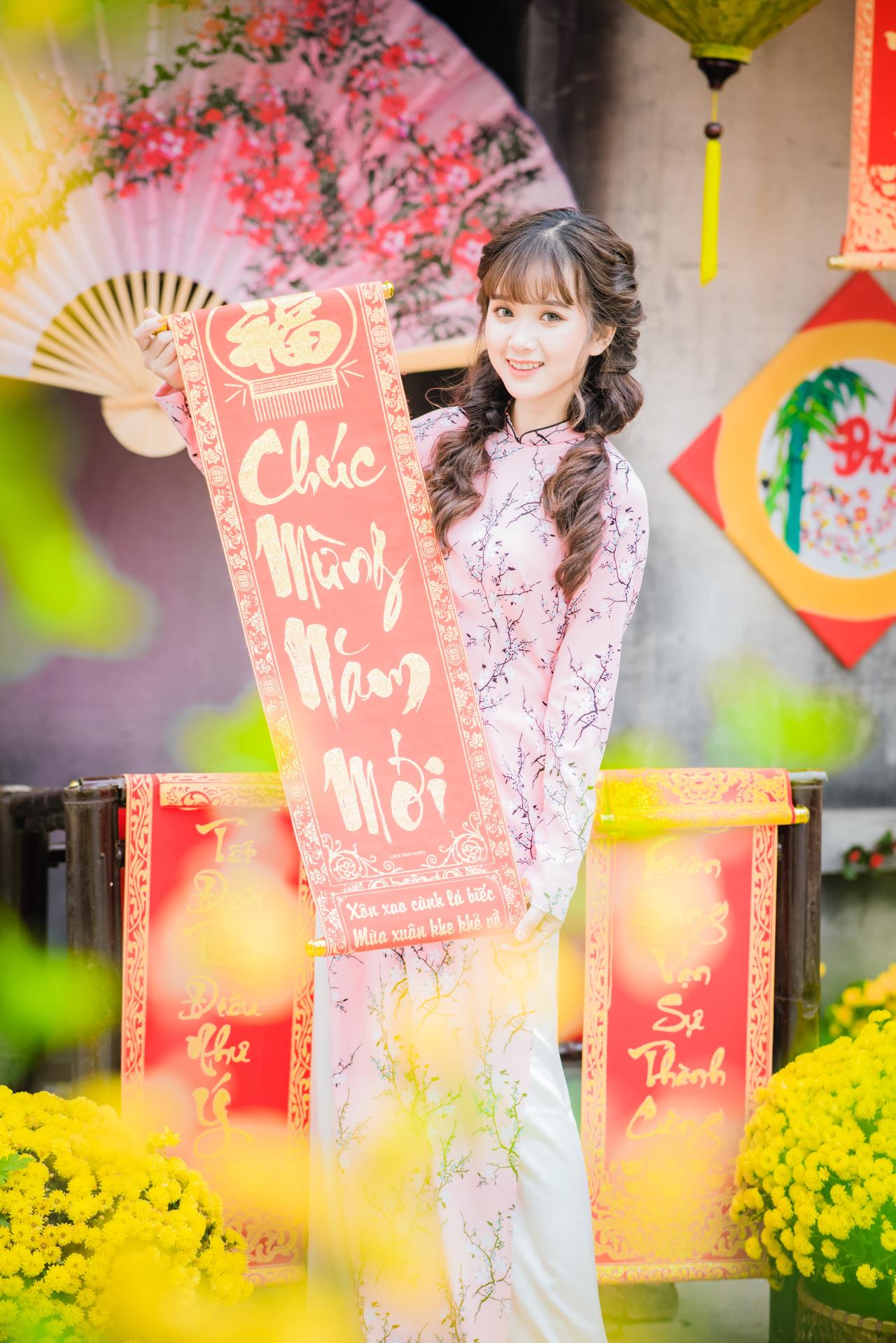棕色双马尾卷发，齐刘海儿，穿着粉色旗袍白色长裤的少女 站姿 全身照 正面照 竖幅 咧嘴露齿微笑 双手拉开越南福字春联 腊梅 黄梅 花篮 粉色梅花扇