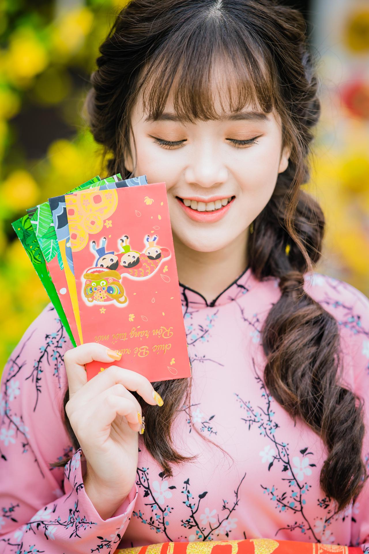 棕色双马尾卷发，齐刘海儿，穿着粉色旗袍白色长裤的少女 站姿 上半身照 正面照 竖幅 低头闭眼 咧嘴露齿微笑 右手拿着彩色幸运签贺卡