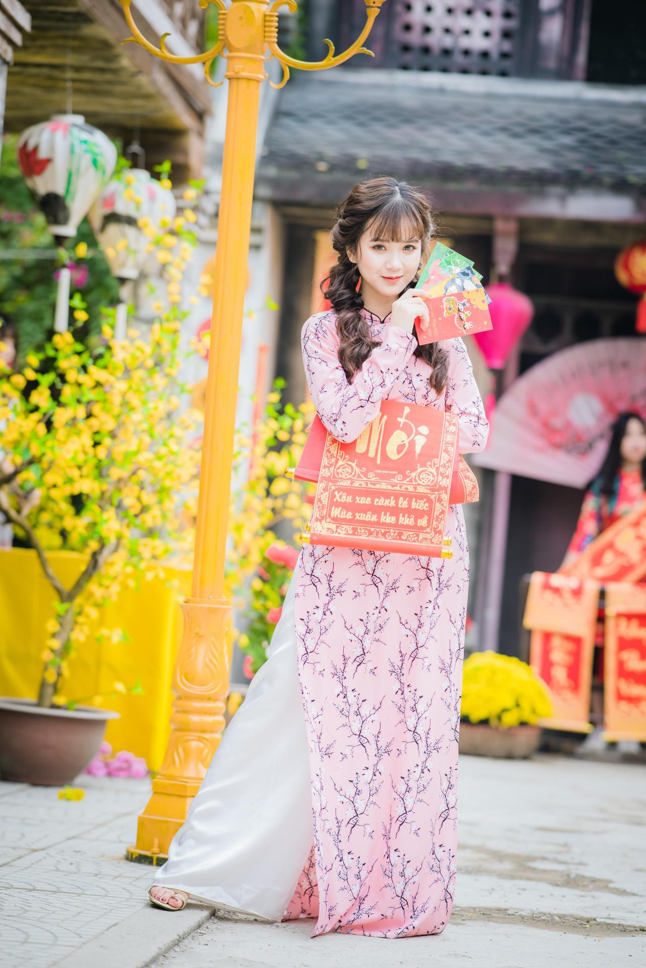 棕色双马尾卷发，齐刘海儿，穿着粉色旗袍白色长裤的少女 站姿 全身照 正面照 竖幅 左手拿着彩色幸运签贺卡 右手拿着越南福字春联 腊梅 黄梅 花盆 橙色灯柱