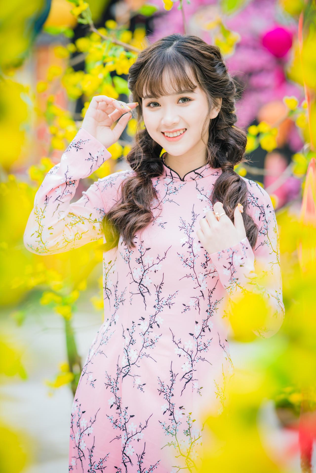 棕色双马尾卷发，齐刘海儿，穿着粉色旗袍的越南少女 站姿 全身照 正面照 竖幅 右手轻抚刘海儿 左手轻抚头发放在胸前 咧嘴露齿微笑 腊梅 黄梅 树