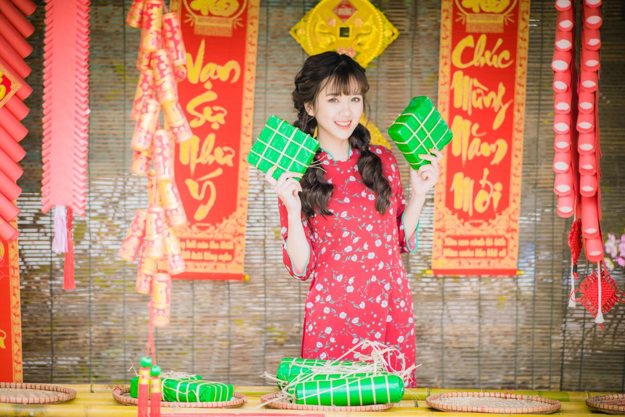 棕色双马尾卷发，齐刘海儿，穿着红色旗袍的越南少女在竹屋前，双手拿着绿色年货包，放在两耳侧 咧嘴露齿微笑 站姿 全身照 正面照 竖幅 中国结 春联 鞭炮