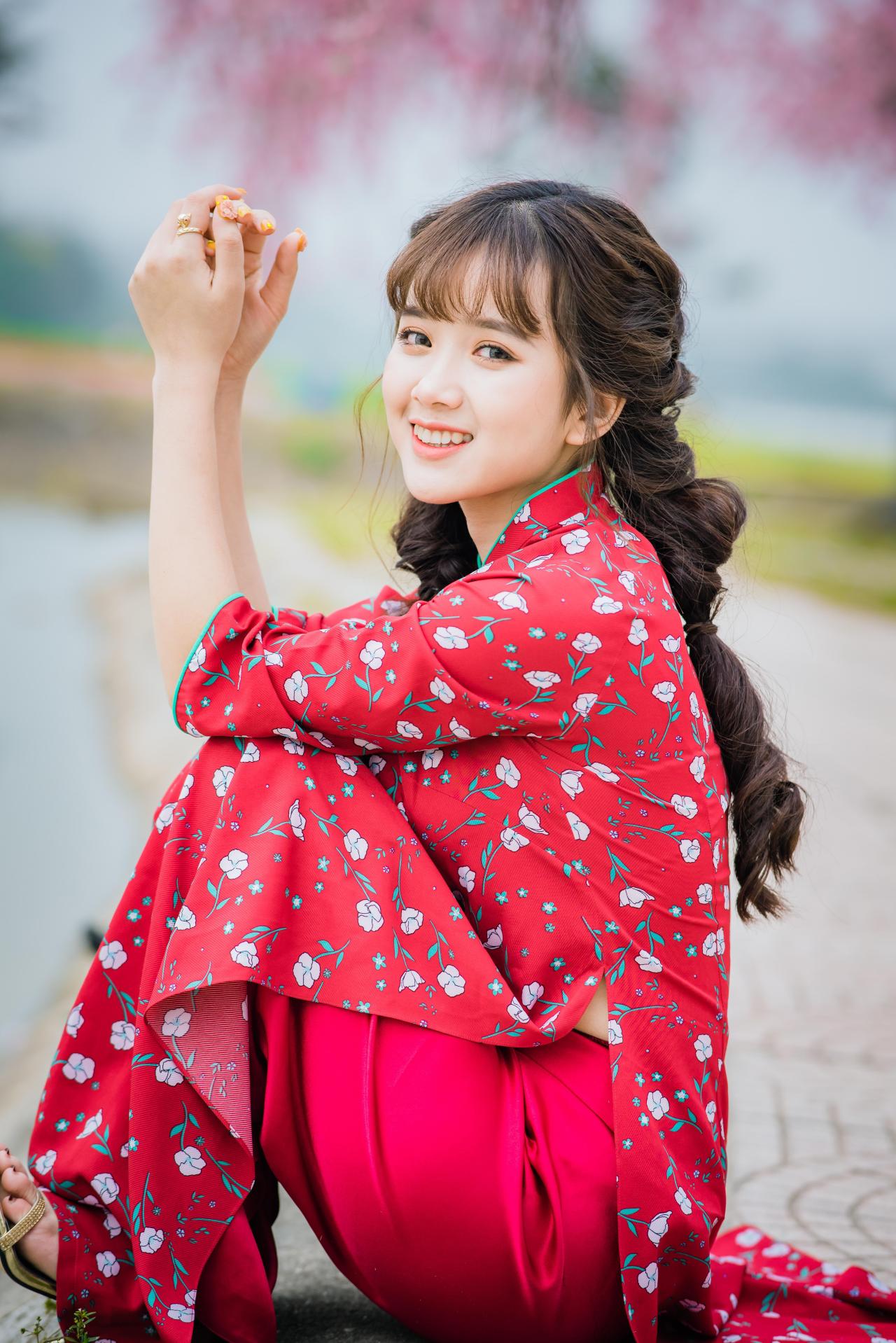 棕色双马尾卷发，齐刘海儿，穿着红色旗袍的越南少女坐在河边 回头看镜头 咧嘴露齿微笑 坐姿 全身照 4/3侧面照 竖幅