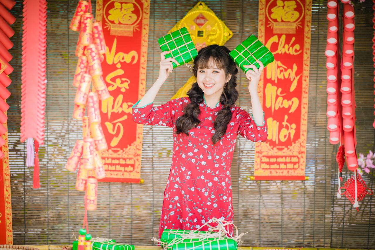 棕色双马尾卷发，齐刘海儿，穿着红色旗袍的越南少女在竹屋前，双手拿着绿色年货包，举过头顶 咧嘴露齿微笑 站姿 全身照 正面照 竖幅 中国结 春联 鞭炮