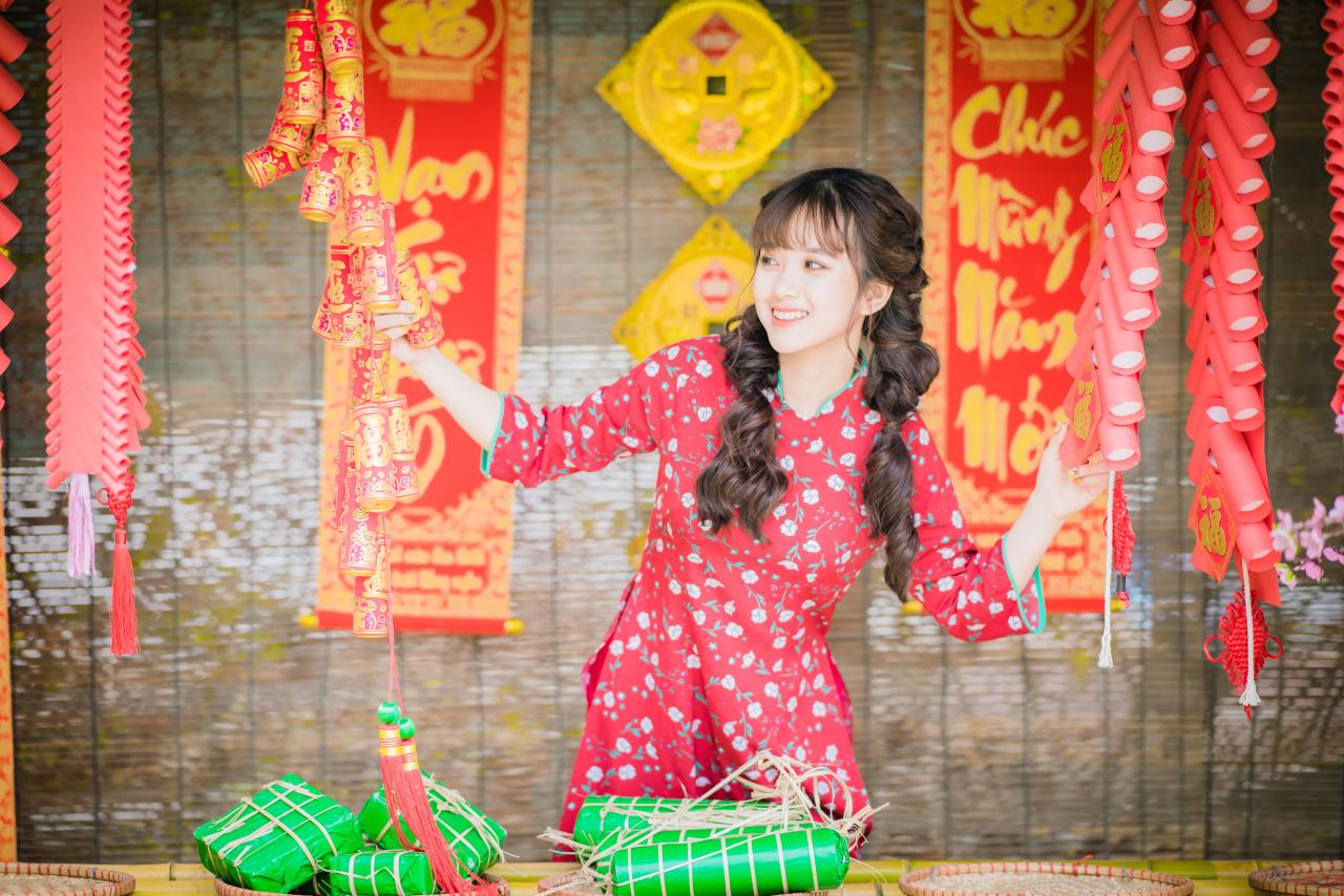 棕色双马尾卷发，齐刘海儿，穿着红色旗袍的越南少女在竹屋前，双手拉开挂着的鞭炮，探出头来 咧嘴露齿微笑 站姿 全身照 正面照 竖幅 中国结 春联